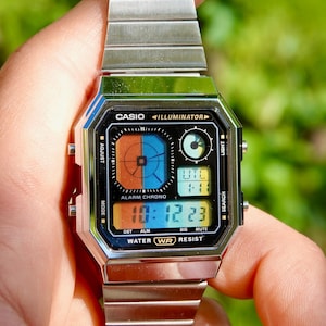 Puede incluir: Un reloj digital Casio plateado con una esfera rectangular y una correa de metal a juego. La esfera del reloj muestra la hora, la alarma y otras funciones. El reloj tiene el texto "CASIO ILLUMINATOR" y "WATER WR RESIST".