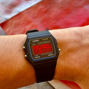 Puede incluir: Un reloj digital Casio F-91W negro con una pantalla roja. El reloj tiene una correa negra y una clasificación de resistencia al agua de WR. El reloj está en la muñeca de una persona.