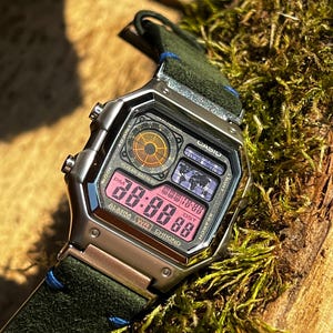 Op de afbeelding: Een zilveren Casio digitaal horloge met een zwart en groen leren bandje. De wijzerplaat van het horloge toont de tijd, datum en alarm. Het horloge heeft een wereldtijdfunctie en een stopwatch.