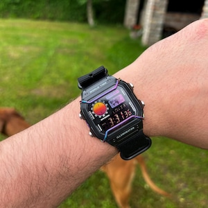 May include: Black digital watch with a rainbow-colored bezel, displaying the time 3:31:36. The watch has a black band and features the text "WORLD TIME" and "ILLUMINATOR". The watch is on a wrist.