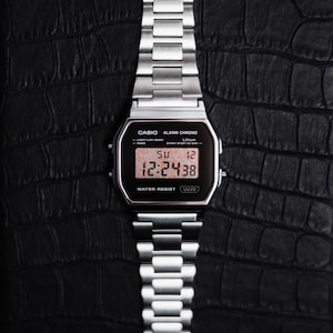 May include: A silver digital watch with a rectangular face and a matching metal band. The watch displays the time as 12:24:38 and the date as SU 12. The watch is labeled "CASIO ALARM CHRONO" and "WATER RESIST".