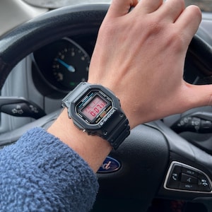 May include: A black G-Shock watch with a digital display showing the time, date, and day of the week. The watch has a black resin strap and a black stainless steel case.