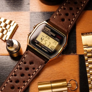 Może przedstawiać: Złoty zegarek cyfrowy Casio z brązowym skórzanym paskiem z perforacjami. Zegarek pokazuje godzinę 4:57:10. Dodatkowe złote elementy zegarka i mały złoty pojemnik są również widoczne, wszystko na szachownicy.