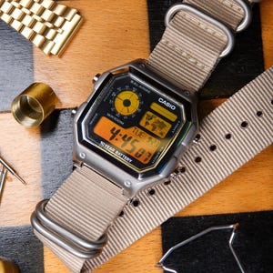 May include: A silver Casio digital watch with a beige nylon strap, displaying the time 4:45:03. The watch is set on a wooden chessboard, with chess pieces and watch components nearby.