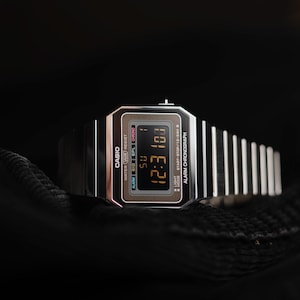 May include: A silver Casio digital watch with a black display showing the time 12:31. The watch features a rectangular face with the Casio logo and function indicators. The watchband is made of silver metal links. The watch is set against a dark background.