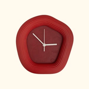 Puede incluir: Un reloj de pared rojo con un marco ondulado único. La esfera del reloj es de un tono rojo más oscuro con manecillas blancas y un diseño minimalista. El marco del reloj tiene un aspecto texturizado y acanalado.