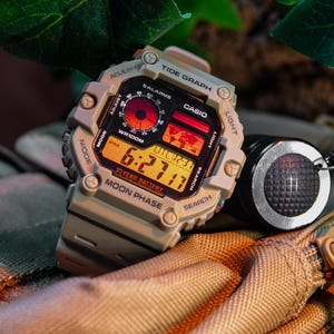 May include: A tan Casio watch with a digital display and multiple functions, including a tide graph, moon phase, and 10-year battery. The watch has a black face with orange and red accents.