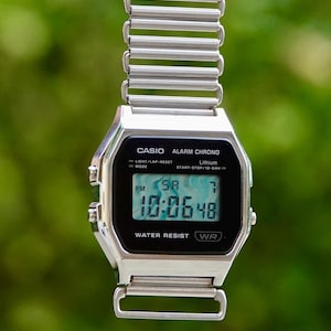 Puede incluir: Un reloj digital Casio plateado con una pantalla negra que muestra la hora. El reloj tiene una esfera rectangular y una correa de metal. El texto "CASIO ALARM CHRONO" es visible en la esfera. El reloj es resistente al agua.
