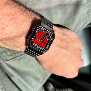 Puede incluir: Un reloj de pulsera Casio Illuminator negro con una pantalla digital roja. El reloj tiene una correa de metal negro y una cara rectangular.