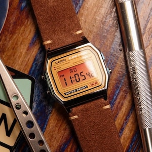 May include: A Casio digital watch with a gold face and black frame, displaying the time. It has a brown suede strap with cream stitching. The watch is water-resistant and has alarm and chrono functions.