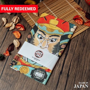 Op de afbeelding: Een kleurrijk bandana met een Japanse krijger met een woeste uitdrukking. Het bandana is gevouwen en vastgebonden met een wit lint waarop "G-SHOCK 40th Anniversary Since 1983" staat. Een zwart digitaal horloge is zichtbaar aan de onderkant van het bandana. Het bandana ligt op een houten oppervlak met rode en bruine decoratieve bloemen.