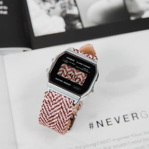 May include: A silver Casio digital watch with a red and white patterned strap. The watch face shows the time. The watch is on a white surface with text and the hashtag #NEVERG.