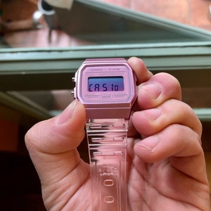 Puede incluir: Un reloj digital Casio F-91W rosa con una banda de metal plateado. La esfera del reloj muestra la hora y la palabra "CASIO" en azul.