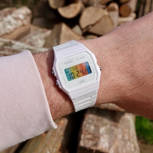 Op de afbeelding: Witte Casio F-91W digitale horloge met de tijd 10:24:20. Het horloge heeft een rechthoekige wijzerplaat met een veelkleurig display en een witte band. Het horloge zit om een pols.