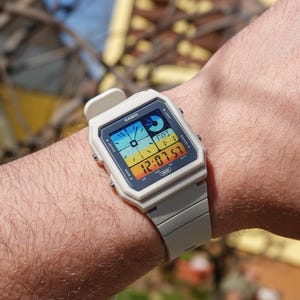 Peut inclure: Une montre numérique Casio blanche avec un écran multicolore. Le cadran de la montre affiche l'heure, la date et le jour de la semaine. La montre est au poignet d'une personne.