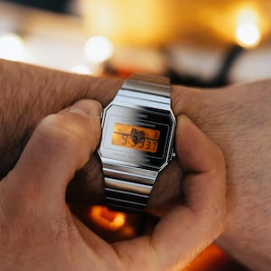 May include: A silver Casio digital watch with a black face and orange digital display. The watch is on a silver metal band and is being worn on a wrist. The watch displays the time 9:33:37.