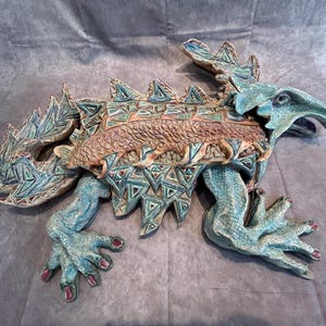 May include: A ceramic lizard sculpture featuring a textured teal and brown body with a pattern of raised triangles along the back. The creature has prominent spikes, a long tail, and four legs with distinct claws tipped in reddish-brown paint.