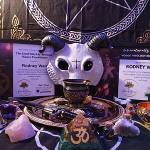May include: A white skull mask with horns, a black and gold chalice, a gold tray with a dagger, a bell, a pink crystal, a small bottle, a gold bowl, and a wooden pyramid with the symbol Om on it. The items are arranged on a black and white patterned surface. The text on the certificates reads "Internationally Certified The Good Witch & Theosophy Master Practitioner Awarded To Rodney Ward" and "Internationally Certified Vodou Theology Master Class Awarded To Rodney Ward".