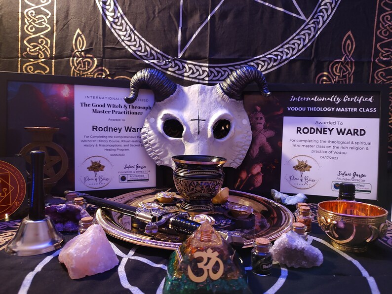 May include: A white skull mask with horns, a black and gold chalice, a gold tray with a dagger, a bell, a pink crystal, a small bottle, a gold bowl, and a wooden pyramid with the symbol Om on it. The items are arranged on a black and white patterned surface. The text on the certificates reads "Internationally Certified The Good Witch & Theosophy Master Practitioner Awarded To Rodney Ward" and "Internationally Certified Vodou Theology Master Class Awarded To Rodney Ward".