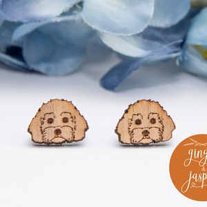 May include: A pair of wooden earrings featuring a laser-cut design of a dog's face. The earrings are a light brown colour and have a natural wood finish. The text "ginger & jasper" is visible in the lower right corner of the image.