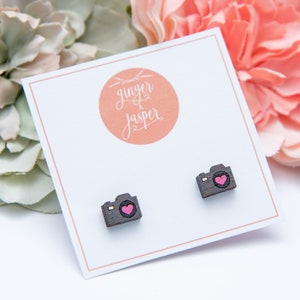 May include: A pair of wooden camera earrings with a pink heart detail. The earrings are displayed on a white card with the text "ginger & jasper" in a circle.