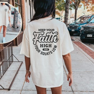 May include: A white oversized t-shirt with a black graphic that says "Keep Your Faith High and Your Squats Low".