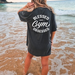 Pu&ograve; includere: Una maglietta grigia scura con il testo "Blessed -and- Gym Obsessed" stampato in bianco sul retro. La maglietta &egrave; indossata da una persona in piedi su una spiaggia sabbiosa.