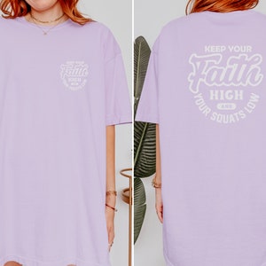 May include: A lavender oversized t-shirt with the text "Keep Your Faith High And Your Squats Low" printed on the back in white. The front of the shirt has a smaller version of the text printed on the left side of the chest.