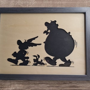May include: A black and white silhouette of a cartoon character with a large belly and two smaller characters walking behind it. The image is framed in a black frame.