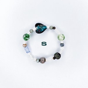 Peut inclure: Un bracelet de perles avec une perle en forme de cœur et d'autres perles vertes, bleues et marron. Le bracelet est enfilé sur un cordon blanc et comprend la lettre "S".
