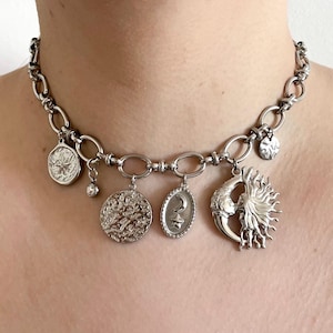 Peut inclure: Collier argenté orné de divers charms célestes. Le collier comprend un pendentif soleil, lune et étoile, ainsi que d'autres breloques circulaires et ovales texturées. La chaîne est composée de maillons ovales interconnectés.
