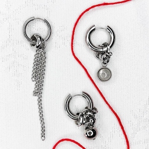 Peut inclure: Trois boucles d'oreilles créoles argentées avec différents breloques. L'une a une chaîne, une autre une pierre transparente, et la troisième un petit charme détaillé avec une perle ronde. Un fil rouge traverse l'image.