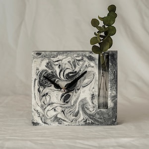 May include: A black and white marbled concrete clock with a small glass vase for flowers. The clock has black hands and a silver center. The vase is empty.