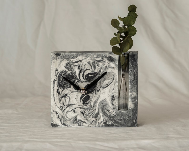 May include: A black and white marbled concrete clock with a small glass vase for flowers. The clock has black hands and a silver center. The vase is empty.