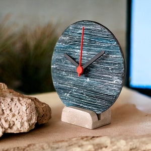 Può includere: Un orologio rotondo, nero e blu con una lancetta dei secondi rossa e lancette delle ore e dei minuti nere. L'orologio è su una base di cemento grigia.