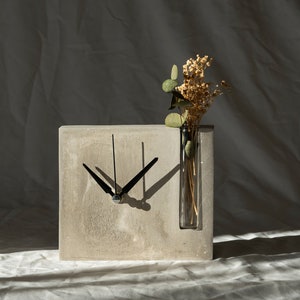 May include: A gray concrete clock with black hands and a clear glass vase with dried flowers inserted into the top. The clock is sitting on a white fabric surface.