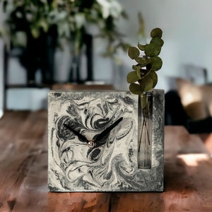 May include: A black and white marbled concrete clock with a glass vase for a single stem of greenery. The clock is on a wooden table.