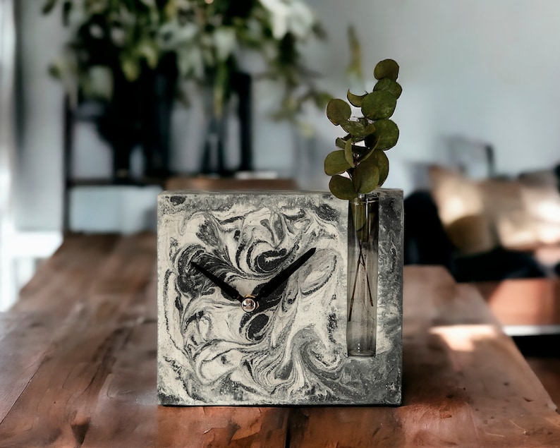 May include: A black and white marbled concrete clock with a glass vase for a single stem of greenery. The clock is on a wooden table.