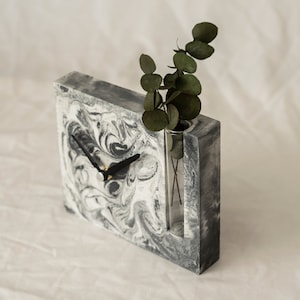 May include: A gray and white marbled concrete clock with a black clock hand and a glass vase insert. A sprig of green leaves is in the vase.