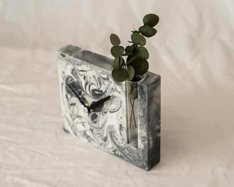 May include: A gray and white marbled concrete clock with a black clock hand and a glass vase insert. A sprig of green leaves is in the vase.