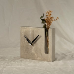 May include: A gray concrete clock with black hands and a clear glass vase for flowers. The clock is sitting on a white surface.