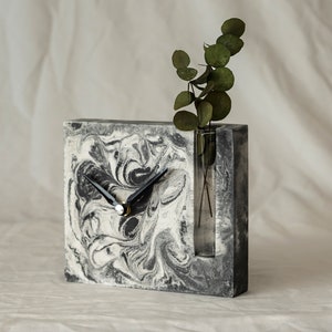 May include: A gray and white marbled concrete clock with black hands and a clear glass vase insert. The vase is filled with green leaves.