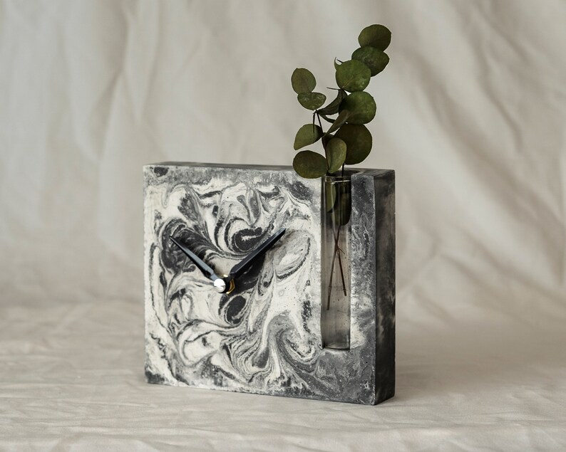 May include: A gray and white marbled concrete clock with black hands and a clear glass vase insert. The vase is filled with green leaves.