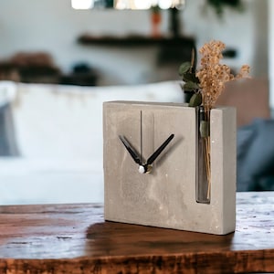 May include: A gray concrete clock with a black clock face and hands. The clock has a built-in glass vase for dried flowers.