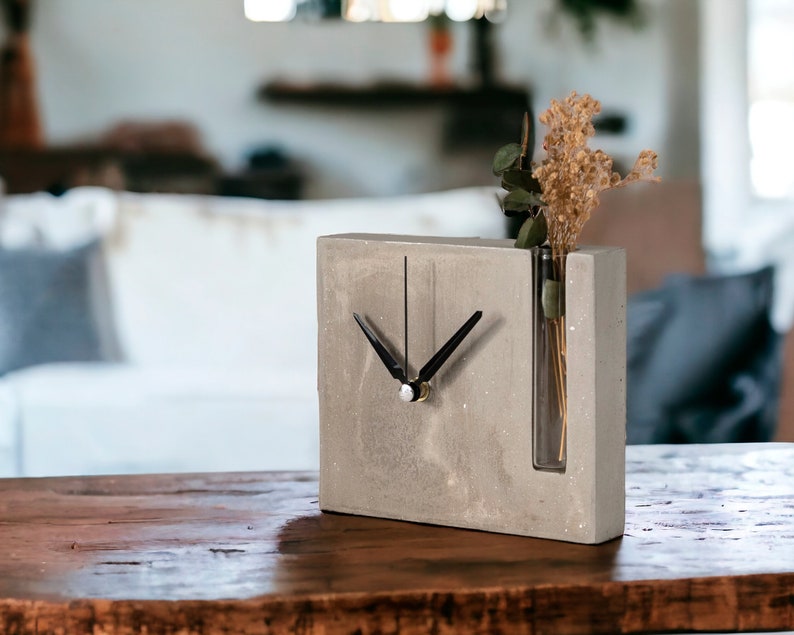 May include: A gray concrete clock with a black clock face and hands. The clock has a built-in glass vase for dried flowers.