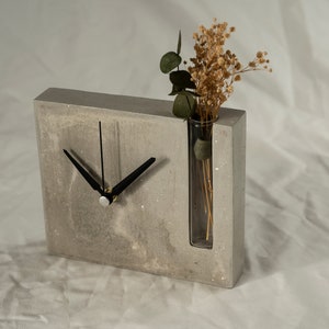 May include: A gray concrete clock with black hands and a glass vase for dried flowers. The clock is on a white surface.