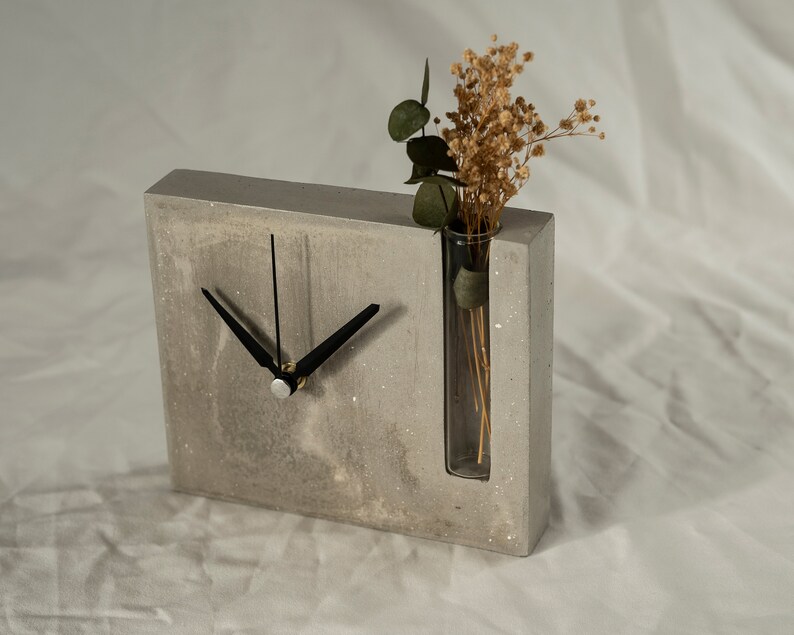 May include: A gray concrete clock with black hands and a glass vase for dried flowers. The clock is on a white surface.