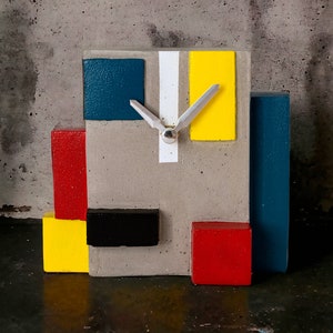Könnte beinhalten: Eine Betonuhr mit geometrischem Design. Das Zifferblatt der Uhr ist grau mit weißen, gelben, roten und blauen Akzenten. Die Zeiger der Uhr sind silberfarben.