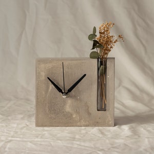 May include: A gray concrete clock with a black clock face and hands. The clock has a built-in glass vase with dried flowers.