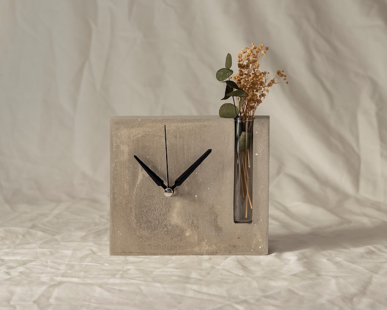 May include: A gray concrete clock with a black clock face and hands. The clock has a built-in glass vase with dried flowers.
